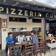 Outdoor seating area of Pizzeria Conza with three men at a long wooden table looking at menus outside the restaurant signage in view.